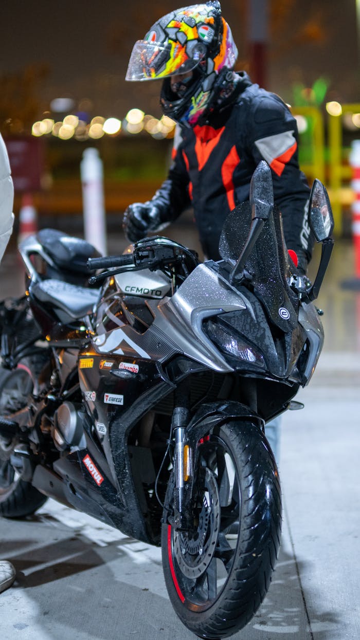 A motorcycle rider in protective gear stands beside a wet sport bike at night.
